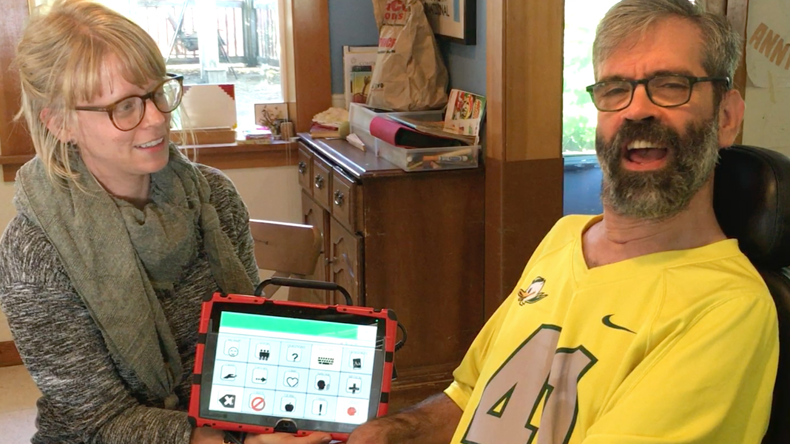 A woman holds a communications device and a man in glasses and a yellow shirt smiles at the camera
