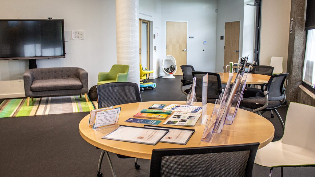 A large room with tables, chairs, a couch, a run, and a large flatscreen TV. The room follows Universal Design principles, including high contrast between the floor and tables. There are many brochures and booklets on one of the tables