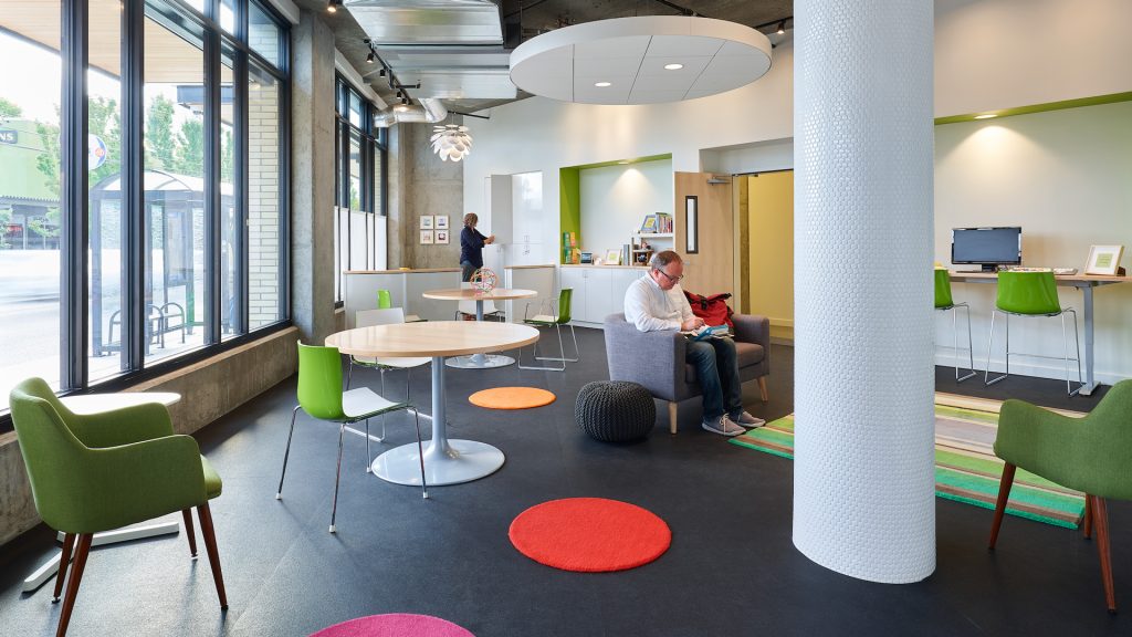 bright room with high contrast floor, tables and chairs. a man sits and reads while a woman in the background opens a cabinet