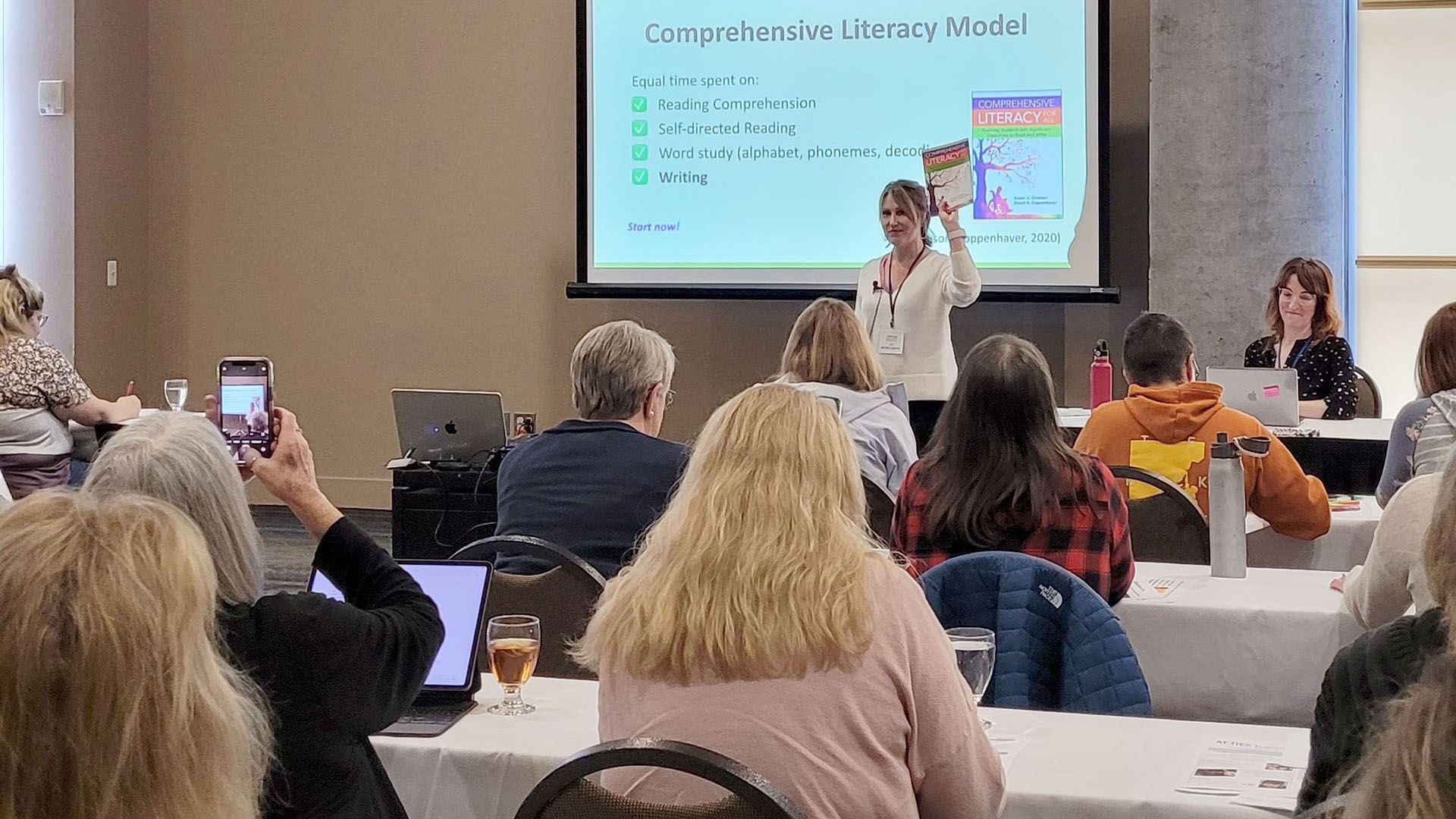 A woman stands in front of an audience with a projections screen behind her that says "Comprehensive Literacy Model"