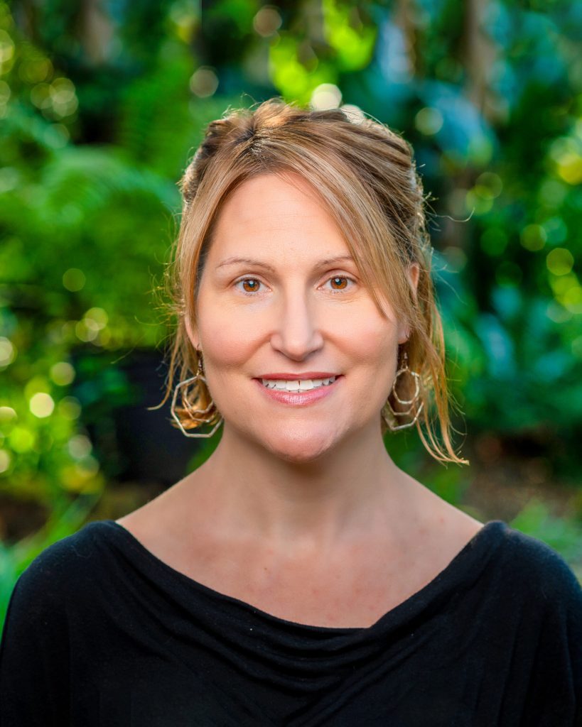 Carrie Luse has blond brown hair and a kind smile. She wears swingy earrings and a black top.