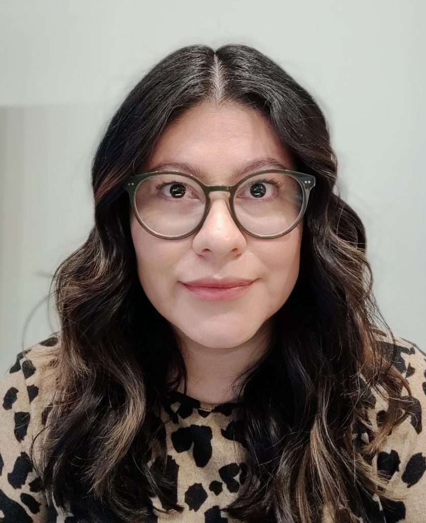 Cynthia Castaneda has long wavy dark hair and wears round glasses. She wears a top with leopard print on it.