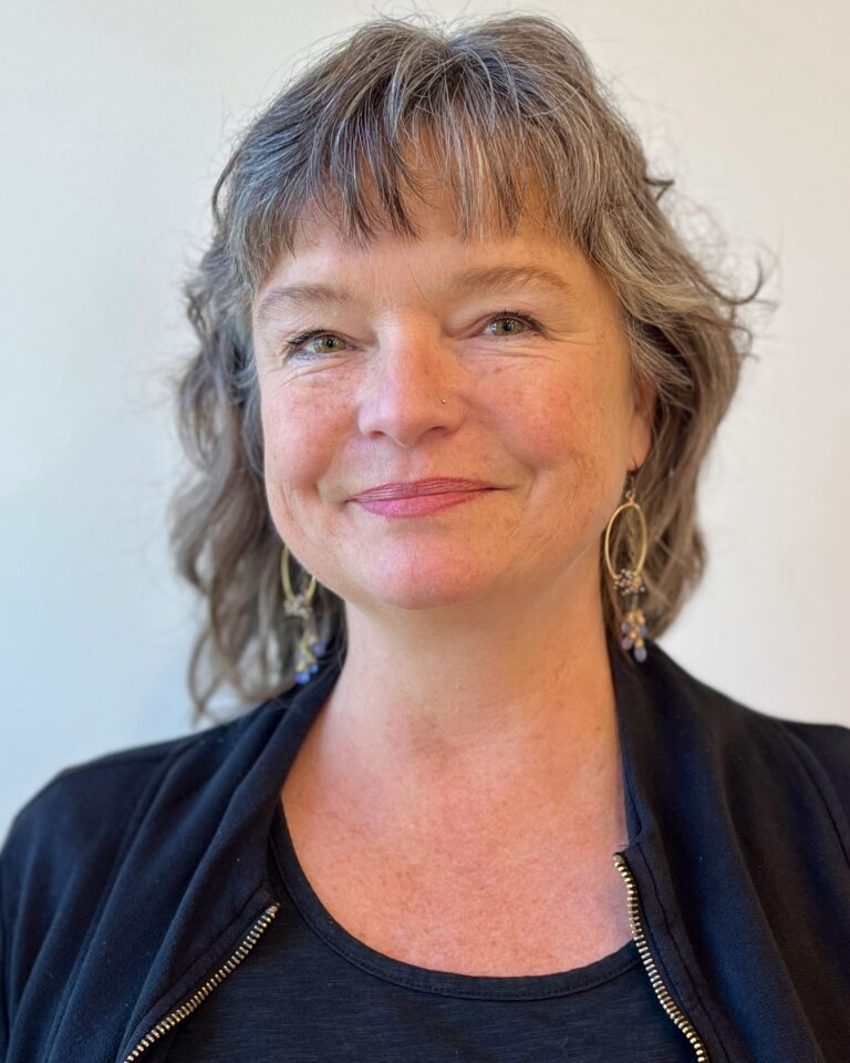 Kim Elliott has dark hair streaked with grey and a warm smile. She wears a black jacket over a black t-shirt.