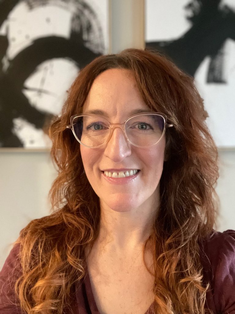 Noelle Birky has long auburn hair and wears glasses. She wears a maroon top.