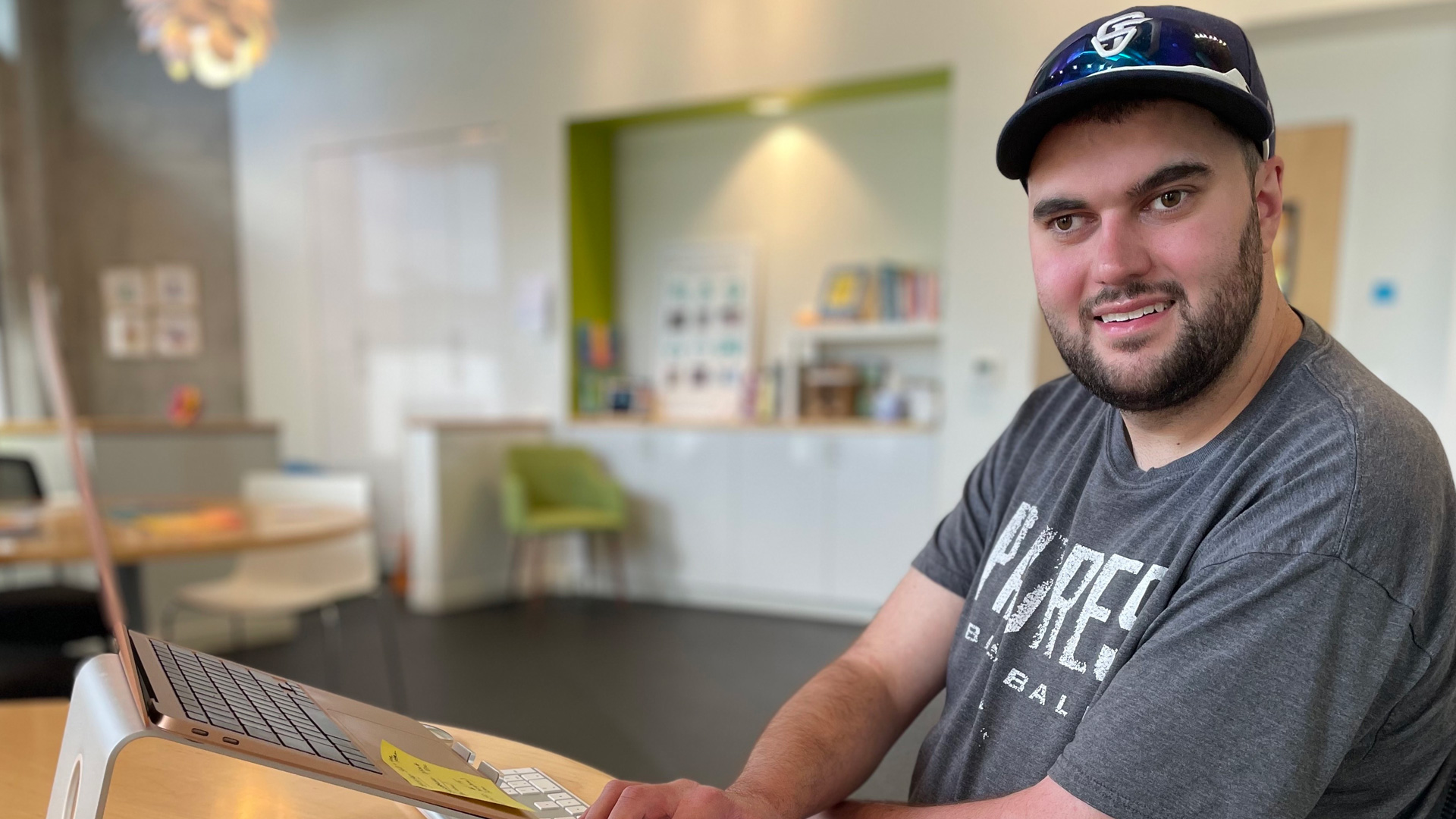 A young man in a basebal hat sits at a table with a laptop on a stand and a keyboard