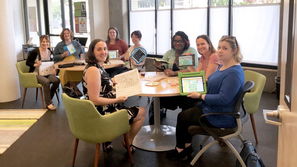 Workshop participants sit around two tables and hold up examples of AAC