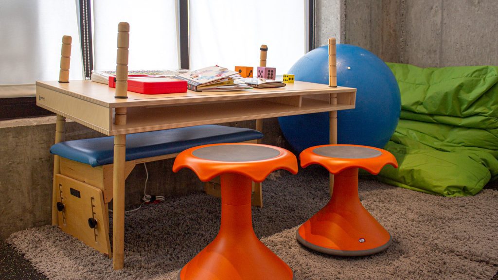 an area for children with orange wobble stools, a green bean bag, a large blue ball, and a table with colorful books on it