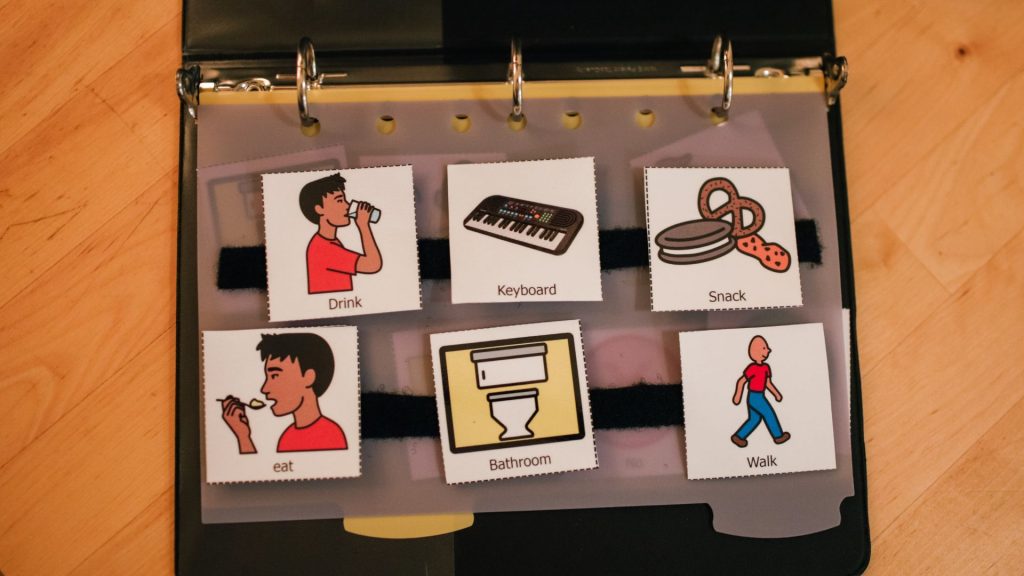 A communication book rest on a table. There are images and words for drink, eat, keyboard, bathroom, snack, and walk.