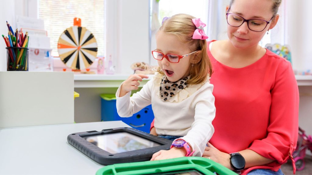 Young girl with red glasses looks at a communication tablet and sits on the lap of a woman in glasses in a colorful office