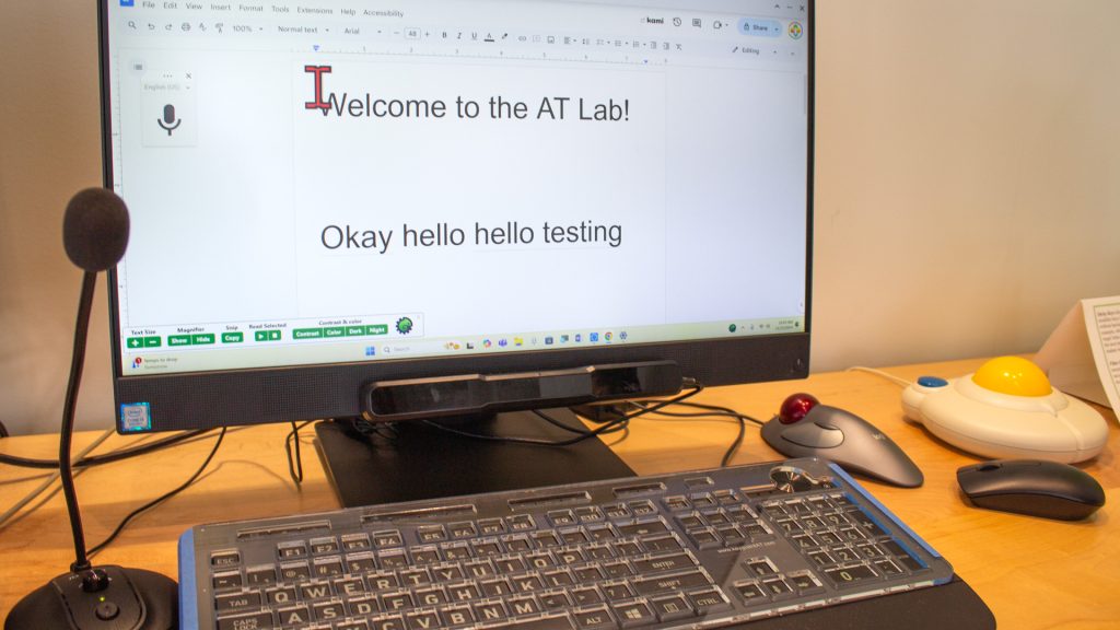 computer screen says "Welcome to the AT Lab! Okay hello hello testing." below the computer screen is a keyboard with a keyguard and different mice