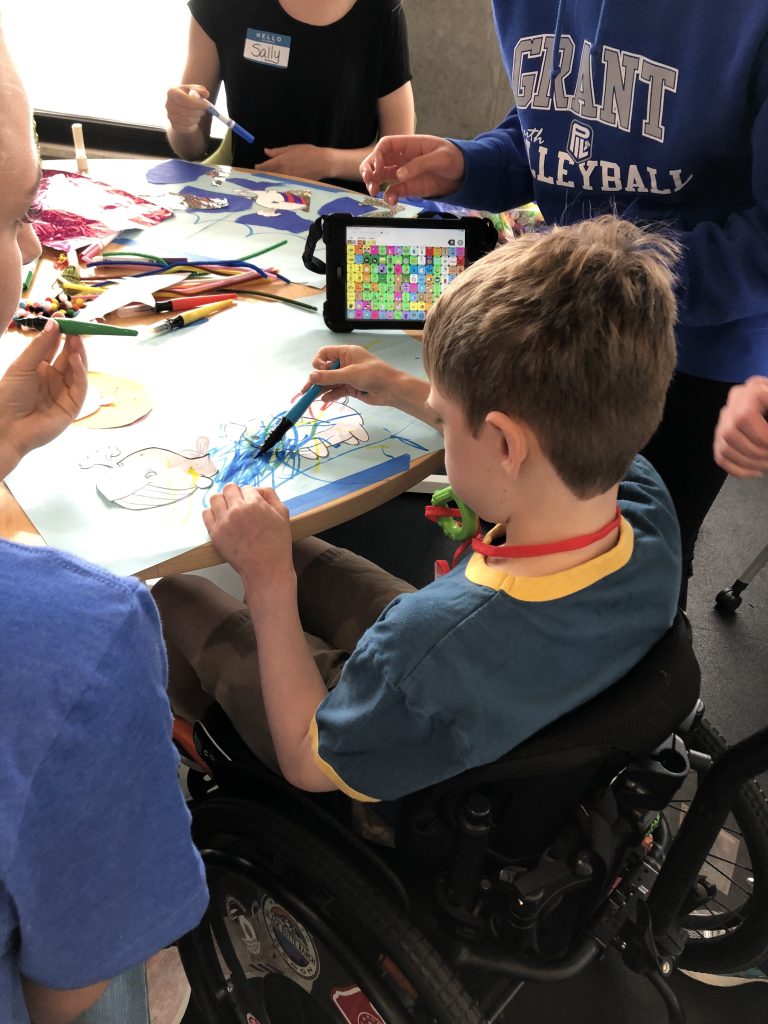 Child draws with other people. There is a communication device on the table.