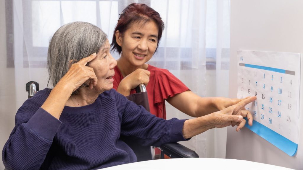 An older woman with her supporter professional looks at a wall calendar.