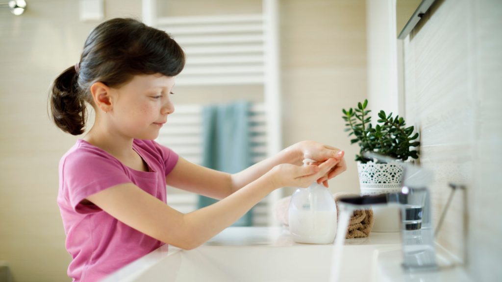 Child in pink shirt washes hands in a light filled bathroom.